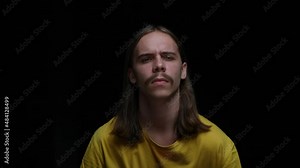 Portrait of a young guy with interest looking into the camera and showing a call me gesture. Male model with long hair and mustache posing on a black background in the studio. Close up. Slow motion.