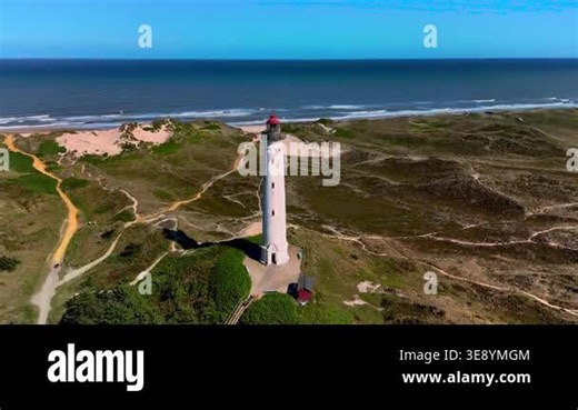 An aerial view of a tall white lighthouse standing on a grassy dune overlooking the blue ocean Stock Video Footage - Alamy
