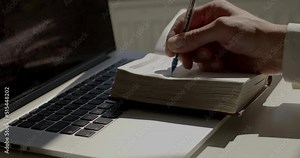 Man hand in suit makes notes with pen in a paper diary at laptop, closeup. Skill of writing by hands, penmanship. Recording thoughts and ideas in a notebook.