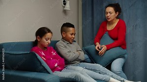 Upset diverse multiethnic children sitting on sofa while annoyed mother scolding kids at home. Judicial woman lecturing preteen boy and girl indoors. Complicated relationships, family conflict