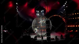 Three acrobats ride motorcycles in a large metal round cage in a circus with colorful lighting. Performance of circus performers on a motorcycle. Dangerous motorcycle stunts. Acrobats on motorcycles.