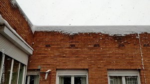 View from a window of snow falling quite heavily. There is a building quite close right in front and two windows and the roof top can be seen.