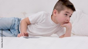 little boy 7 years old lies on bed in hands with tablet online learning interesting-movie mobile games applications I am alone at home spend time on the Internet social networks children and computer