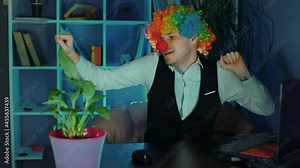 A young man in the image of a clown dances in the workplace, sitting at a computer in the office. The concept of a clown at work