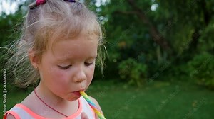 Little child girl eat lick suck a sweet colored caramel candy stick in green garden