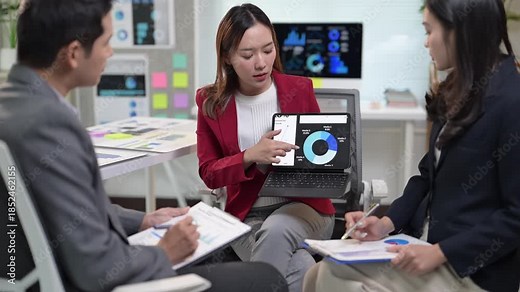A woman is pointing at a pie chart on a laptop. She is sitting in front of a man and another woman