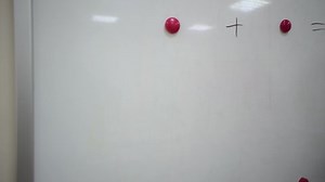 Female teacher stands at whiteboard and explains rules of addition in elementary school. Caucasian schoolmaster in glasses makes mathematical examples using colored magnets and marker.