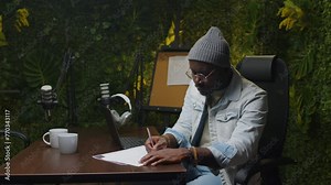 Young African American blogger using laptop and writing podcast script on paper while preparing for recording in audio studio