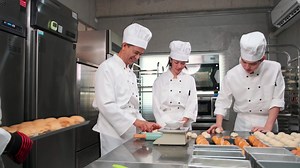 Download Multiracial professional gourmet team, four chefs in white cook uniforms and aprons knead pastry dough and eggs, prepare bread, and bakery food, baking in oven at stainless steel restaurant kitchen. for free