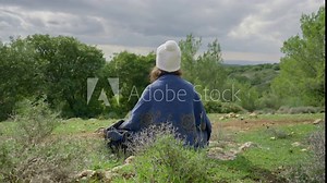 Young hippie free spirit woman sitting in nature and meditates peacefully. Breathing deep and performing spiritual ceremony ritual female sits with back to camera dressed with bohemian style clothes