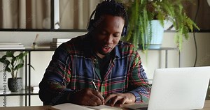 Happy young mixed race biracial african man in headphones listening educational lecture online, communicating with teacher mentor online using video call zoom web camera application, taking notes. Stock Video