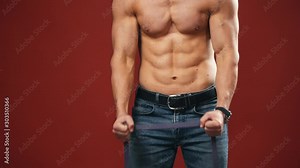 Man's strong body. Shirtless muscular body of a man in jeans doing exercises. Trained sportsman in the studio. Close-up.