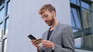 Happy business man holding bank card, entering cvv code or payment information in mobile phone internet shopping application, feeling amazed of purchasing goods or e-banking services online.