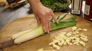 Chopping leek, Garlic cloves, Knife control. Close-up hands using knife to chop leek on wooden cutting board, surrounded by sliced garlic and fresh vegetables.