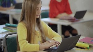 Cute blonde teen girl doing her class work typing on a laptop computer in a classroom during lesson.