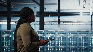 Data center technician monitors data center metrics on tablet to enhance equipment operational effectiveness. Employee walks in server hub, using analytics on device to predict performance, camera B