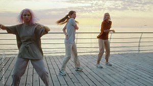 Young dancing teenager girls enjoying funky hip-hop moves. Modern women performing freestyle dance together on sea embankment at sunrise. Slow motion.