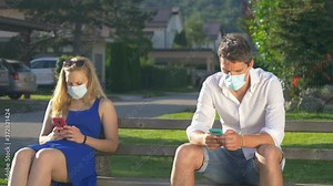CLOSE UP: Two young strangers wear surgical facemasks while sitting on a park bench and texting during the coronavirus pandemic. Two millennials sitting on public bench and wearing protective masks.