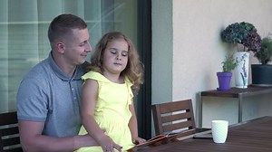 Daddy is kissing his daughter while reading the book at the table. Child listens attentively and leafing through the pages. Father’s day concept