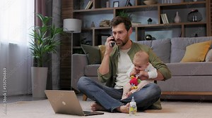Dad is on maternity leave, sitting on a rug in an apartment with a laptop and a baby in his arms. Multitasking father, talking on the phone, working on a laptop and feeding the baby.