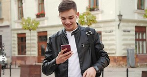 Young Attractive Boy smiles charmingly while Texting Message on his Smartphone. He’s using portable Bluetooth Modern Headphones. He’s casual dressed. Student Lifestyle. Social Network. Apps.