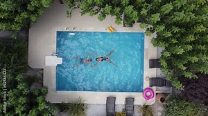 Young couple enjoying summer by pool - both swimming in pool in supine position - drone hovering over pool in bird's eye view
