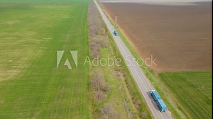 Two large trucks with cargo trailers for loading grain wheat and barley driving along paved road. Concept of goods delivery by automobile. Long endless highway between agronomic fields with crops