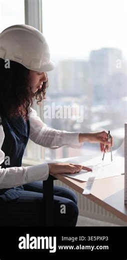 A female architect wearing a hard hat uses a compass and ruler to review architectural plans at her desk with a city view Stock Video Footage - Alamy