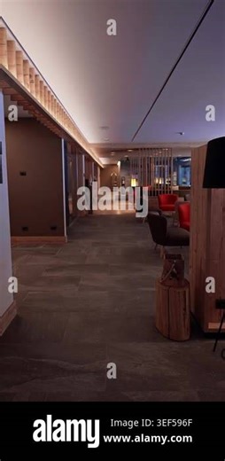 Vertical walkthrough shows an empty Alpine hotel lounge and corridor with slate tile floor, exposed wooden beams, slatted wood ceiling, orange and gray chairs, and a log stump table Stock Video Footage - Alamy