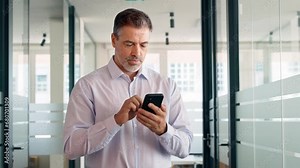 Happy mid aged business man standing in office using cell phone. Mature businessman professional executive manager holding mobile working on smartphone checking applications on cellphone.