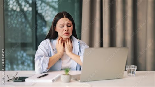 Young woman experiences discomfort, holding throat, indicating sickness. Alone in modern office setting, showing health problems interfering with remote work. Feelings of pain and worry are evident.