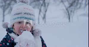 Child eats snow in funny knitted hat outside in winter during snowfall . Small caucasian kid licks snow with tongue.