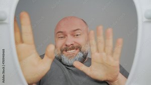 Young man plumber lifts lid of toilet bowl looks inside, dancing and looks funny, having fun at restroom. Bearded guy resting from housekeeping routines. View from toilet bottom. Cleanliness and