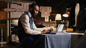 Young fashion designer using laptop in crafting atelier, searching for clothes design inspiration and models. Professional tailor business owner preparing for handcraft process.
