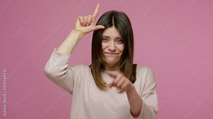 You lose! Brunette young woman making loser gesture, L sign on forehead and pointing to camera, teasing and accusing for defeat, expressing disrespect. indoor studio shot isolated on pink background