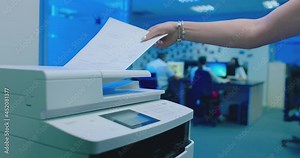 Woman using printer or scanner in office . Office worker use print machine on blurred office background with workers and computers . Hand take papers and press buttons on multifunctional copier .