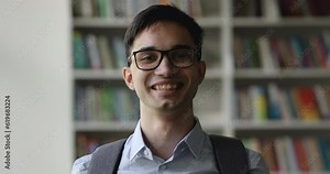 Serious handsome teenage student guy in glasses and backpack posing in library, looking at camera, getting happy, cheerful, positive, laughing, promoting education. Head shot portrait