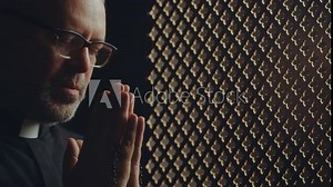 Senior priest wearing glasses holding rosaries in clasped hands and whispering prayer inside of confessional booth. Close-up view