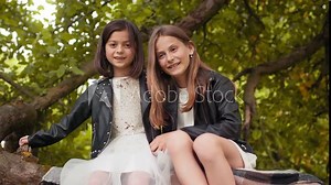 Beautiful little ladies hugging each other while sitting together on tree branch. Two pretty sisters enjoying summertime at green garden. Two sisters sitting together on branch of old tree