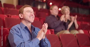 Men and women applaud in a theater with red stadium-style seats. Medium close up with one man in foreground on right. Originally recorded in 4K in slow motion at 60fps with camera on jib arm.