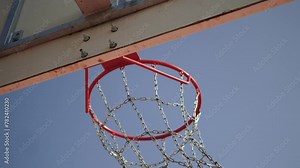 Orange basketball ball hits hoop against blue sky background closeup. Chains of basketball net on basket swing from impact and glisten in sunlight. Pull back during goal in basketball street match.