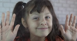 Make faces by the window. A little girl makes funny faces by the glass in the room.