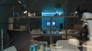 Arc shot of interior of empty experimental biochemical lab at research facility or university at night, with computer, laptop, flasks, vials on tables, and microscope in blurred foreground