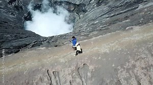 4K Tracking Couple walking by drone on adventure walkway at top of active Volcano with smoke at Mount Bromo (Pananjakan Peak) in Java island, Indonesia. Abstract for freedom life and creative.