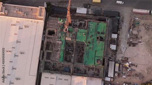 Overhead drone shot of an active construction site with tower crane, rebar grid and green safety netting beside warehouse rooftops, trucks and materials in golden-hour light.