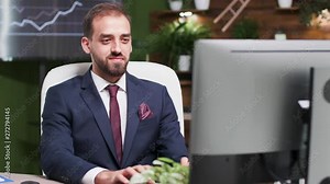 Confident office worker typing on the computer and smiling. He is alone in modern office with creative design. In the background - TV and monitors displaying graphs and sales Stock Video