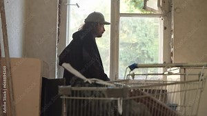 Homeless man is standing in the room of an abandoned building, staring out the window, looking back at the shopping cart of his belongings.