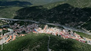 Camera pans over a highway winding through a lush valley in the french pyrenees, revealing the charming village of le perthus nestled amidst the picturesque landscape