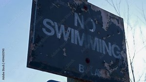 An old no swimming sign. The sun is peeking out from behind it flaring the lens at times. The paint is wrinkling off the surface from being there for so long.