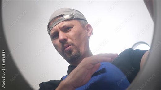 Plumber in a blue worker uniform pinches his nose while looking down, reacting to a bad smell. View from inside a toilet, representing plumbing issues, sewage problems, or cleaning services
